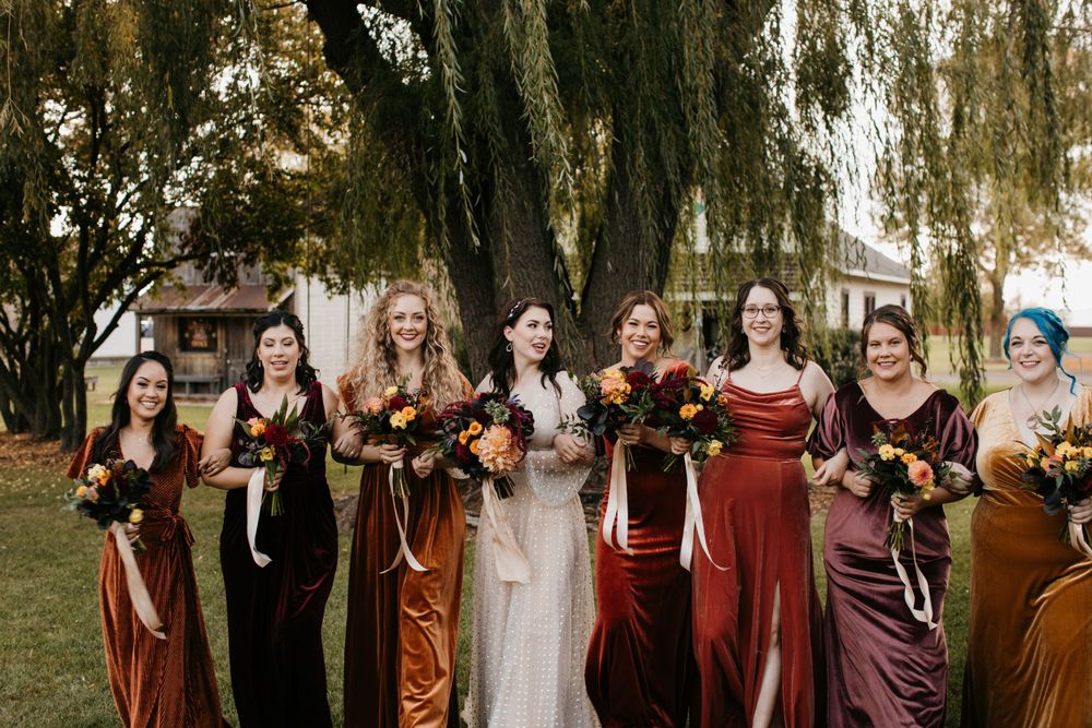 Bride in white gown stands with bridesmaids in velvet fall-toned dresses holding bouquets outdoors.