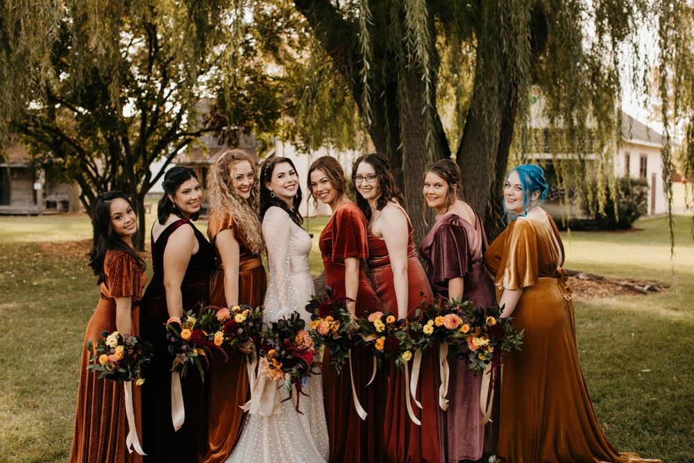Bride and bridesmaids in mismatched velvet autumn dresses pose together smiling under large tree.