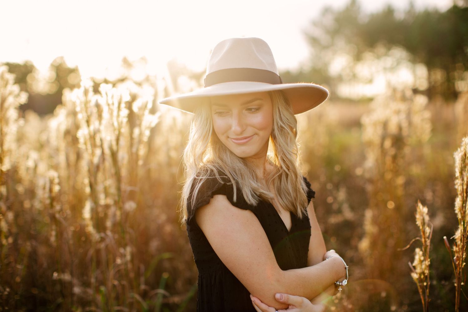 Person in a black dress and wide brim hat poses in a golden field at sunset creating a dreamy summer portrait.