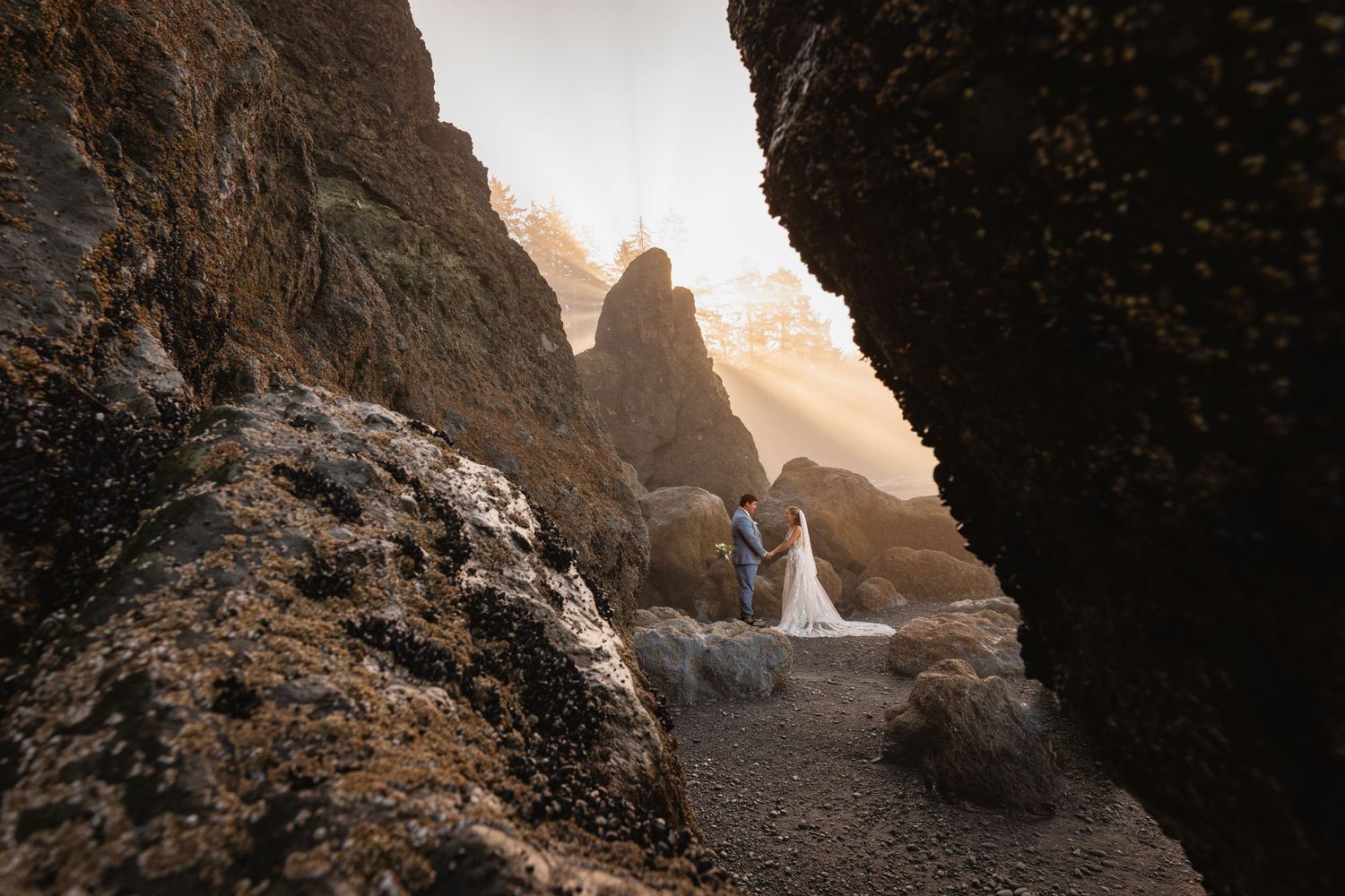 Wedding couple stands framed by dramatic rocky sea cave walls with misty ocean light in the background.