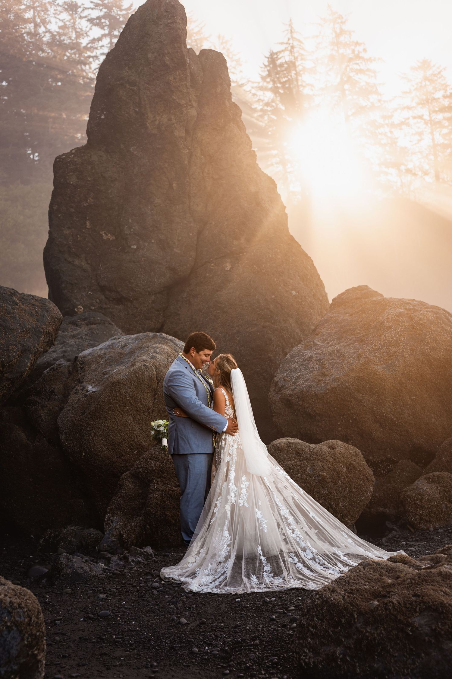 Bride and groom embrace romantically beside a towering rock formation with golden sunbeams piercing through misty trees.