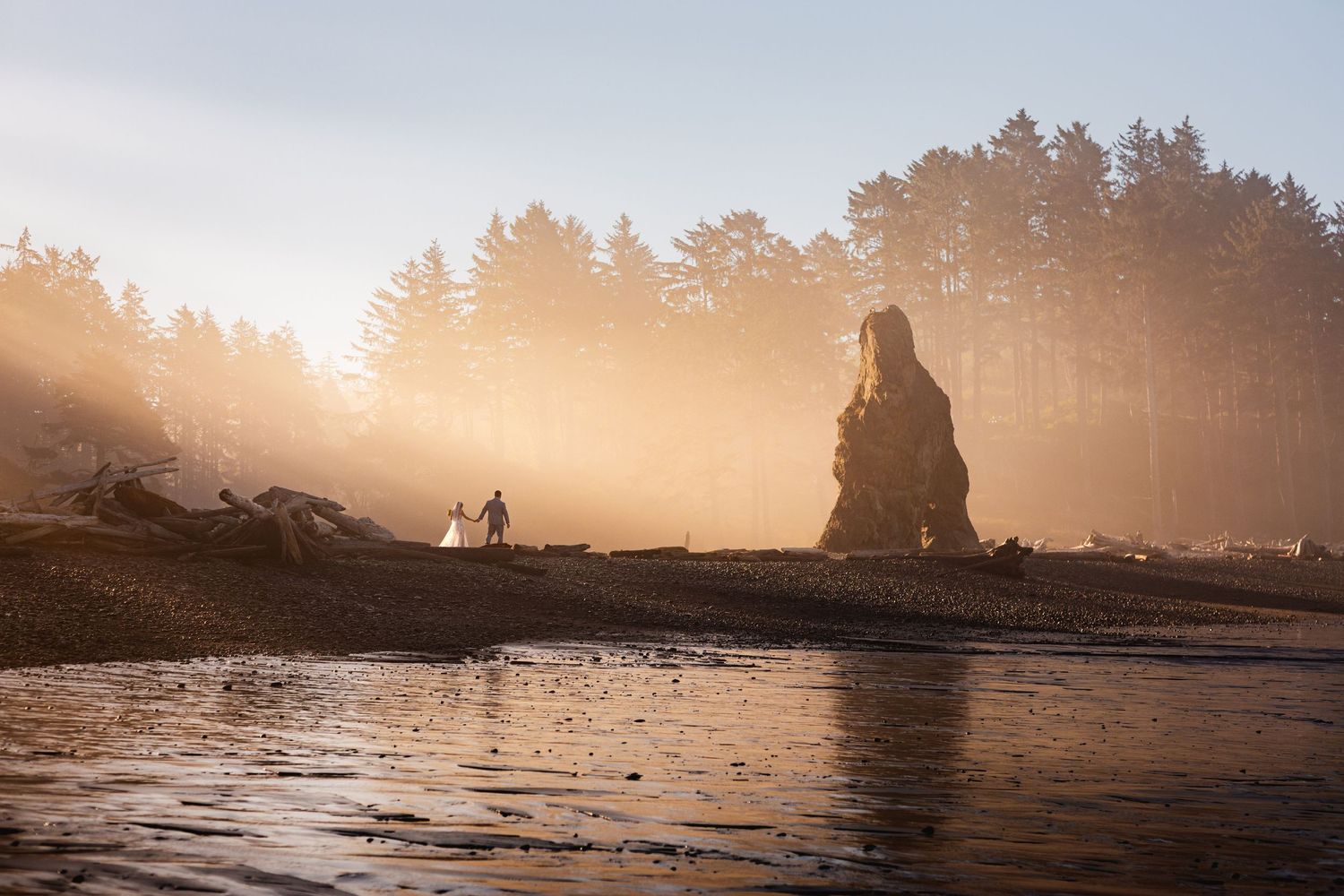 Couple walks along a misty golden beach at sunset with a dramatic sea stack silhouette in the background.
