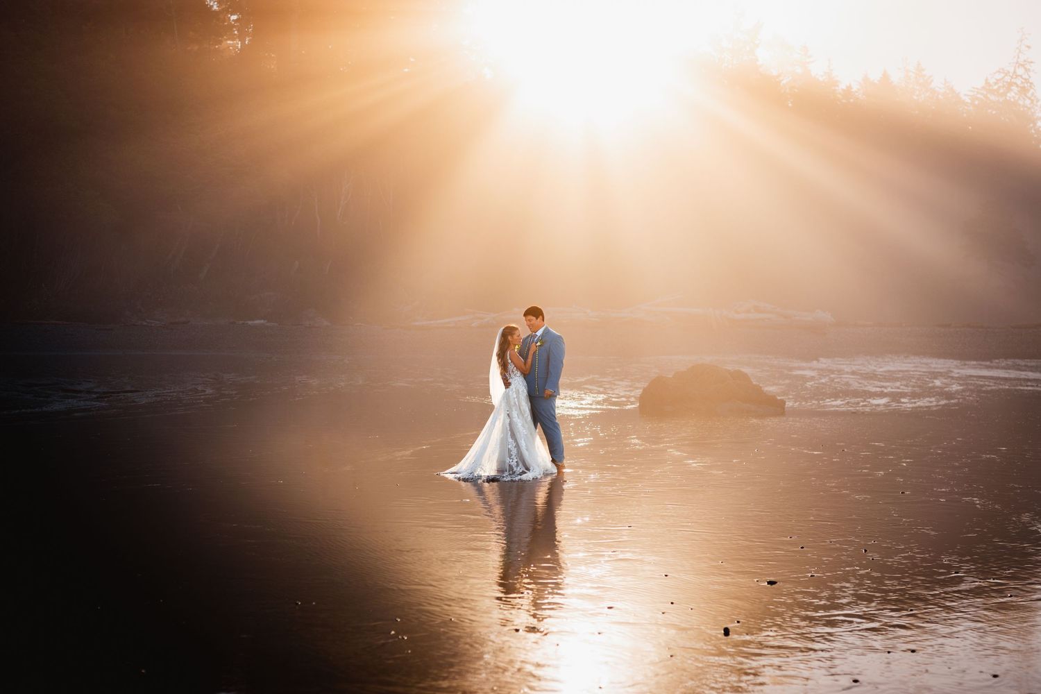 Wedding couple embraces on a misty beach bathed in warm golden sunlight rays during a magical sunset elopement.