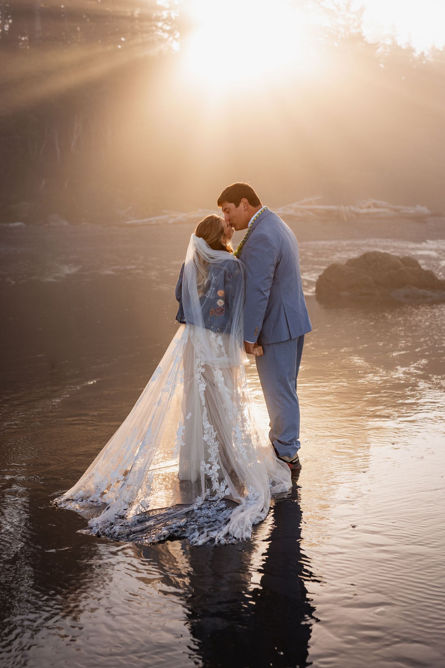 Bride and groom share a romantic kiss standing in shallow water during a stunning golden sunset on the beach.