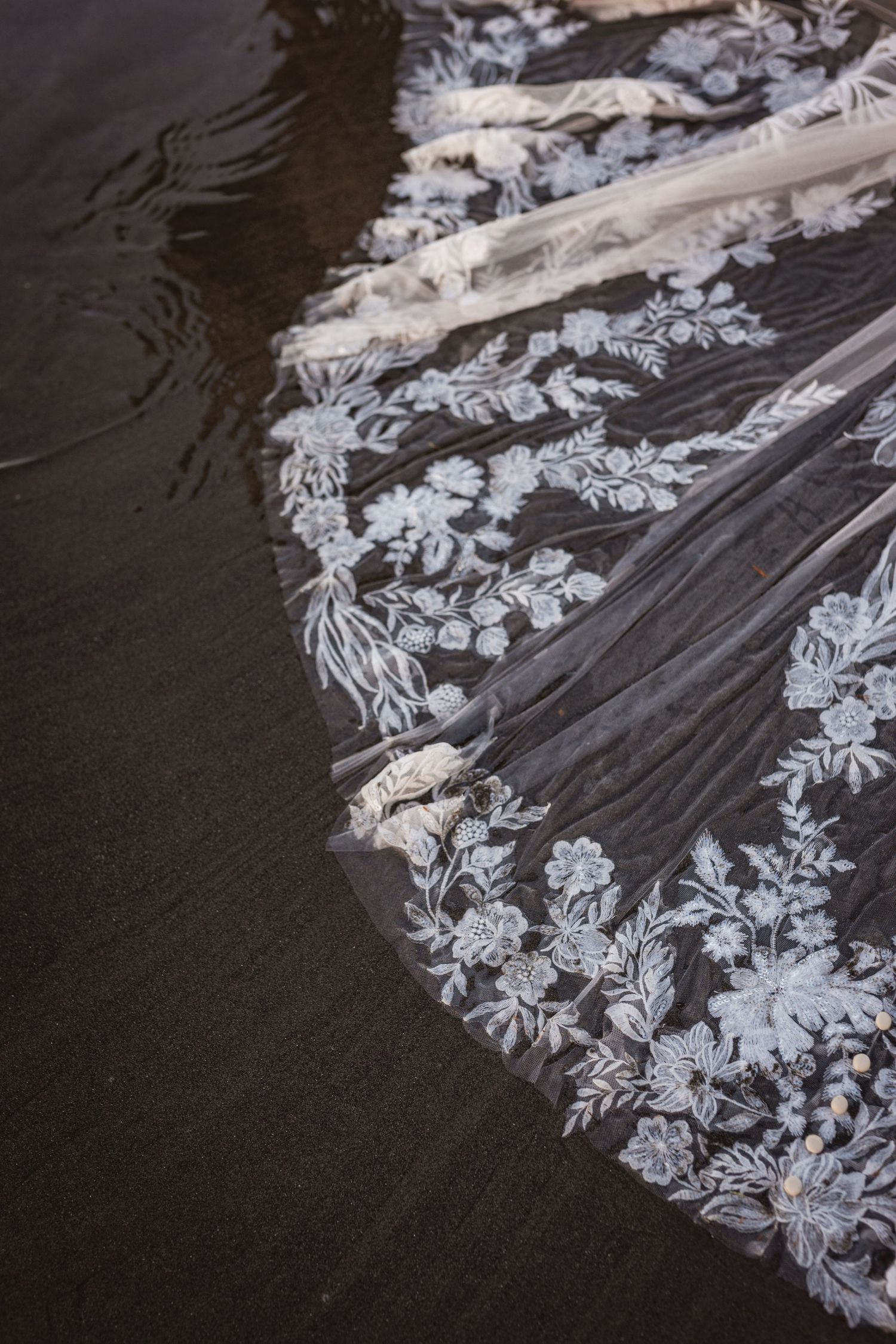 Close-up of intricate floral lace wedding veil trailing across dark wet sand on a beach.