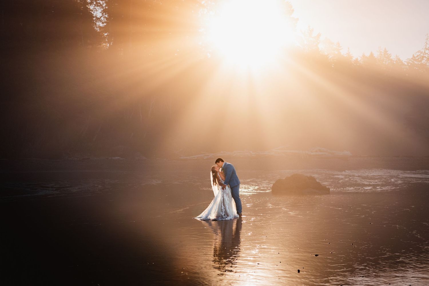 Bride and groom share an intimate moment standing in shallow water surrounded by glowing golden sunset light.