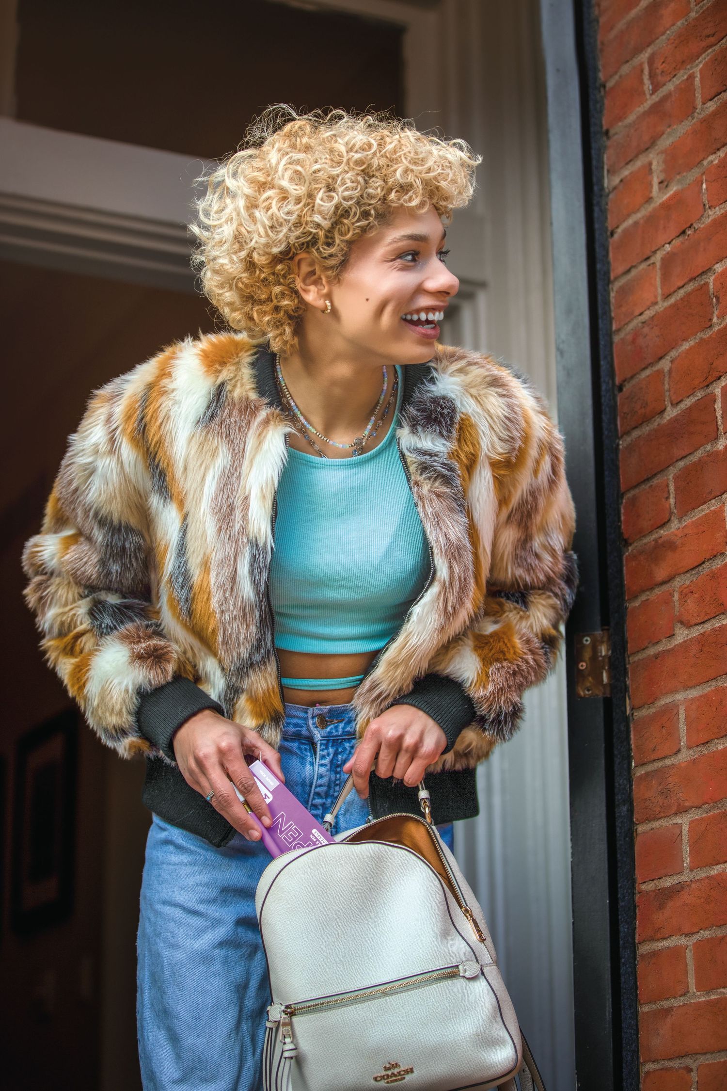 Smiling young woman with curly blonde hair wearing a colorful fur coat and carrying a white backpack.