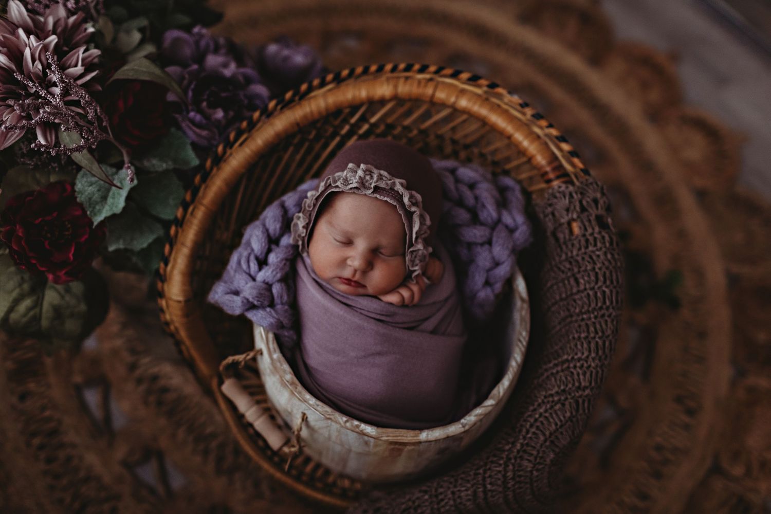 Sleeping newborn in a purple wrap nestled in a round woven basket with delicate lace details.