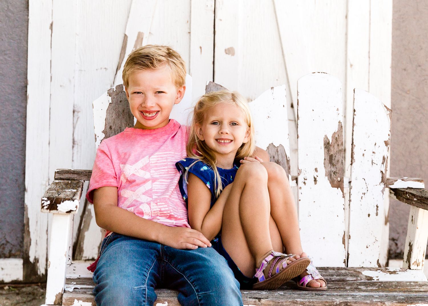 Two young siblings smiling and sitting together on rustic wooden steps outdoors.
