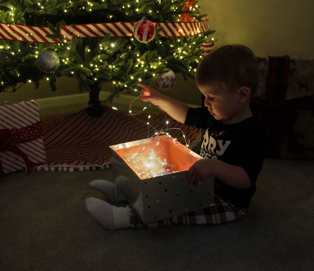 A child in pajamas opens a glowing gift box in front of a lit Christmas tree.