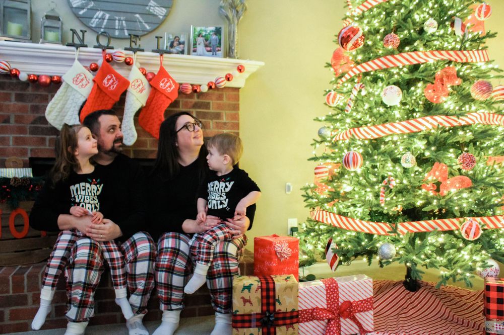 A family in coordinated black and plaid outfits sits by their Christmas tree with stockings hanging above.