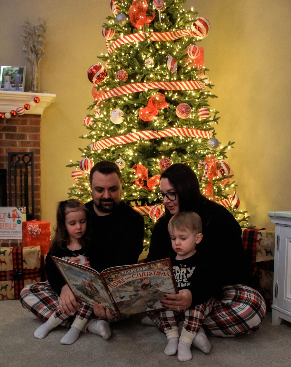 Family in matching pajamas reading together by a decorated Christmas tree with candy cane ribbon.