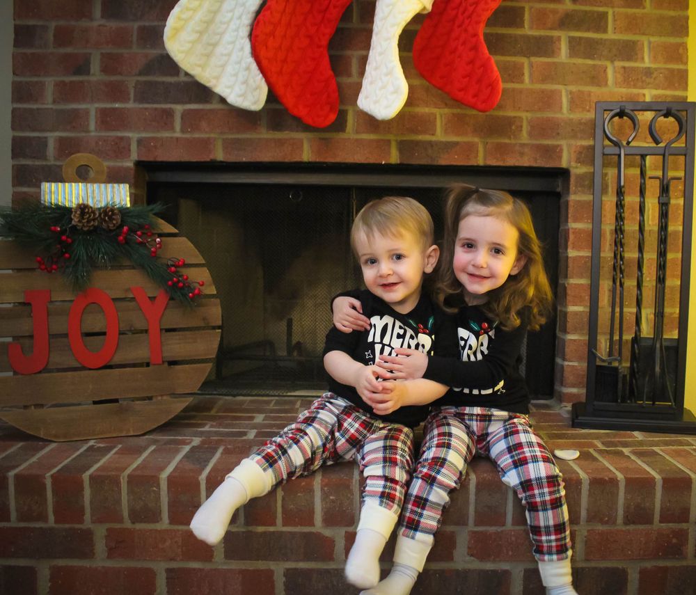 Two children in matching outfits sitting on brick hearth with Christmas stockings hung above.