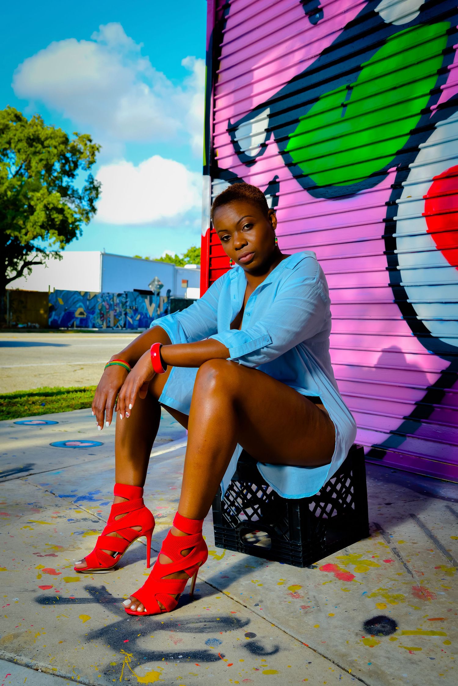 Urban fashion photoshoot featuring model in blue shirt and red heels against colorful graffiti wall.