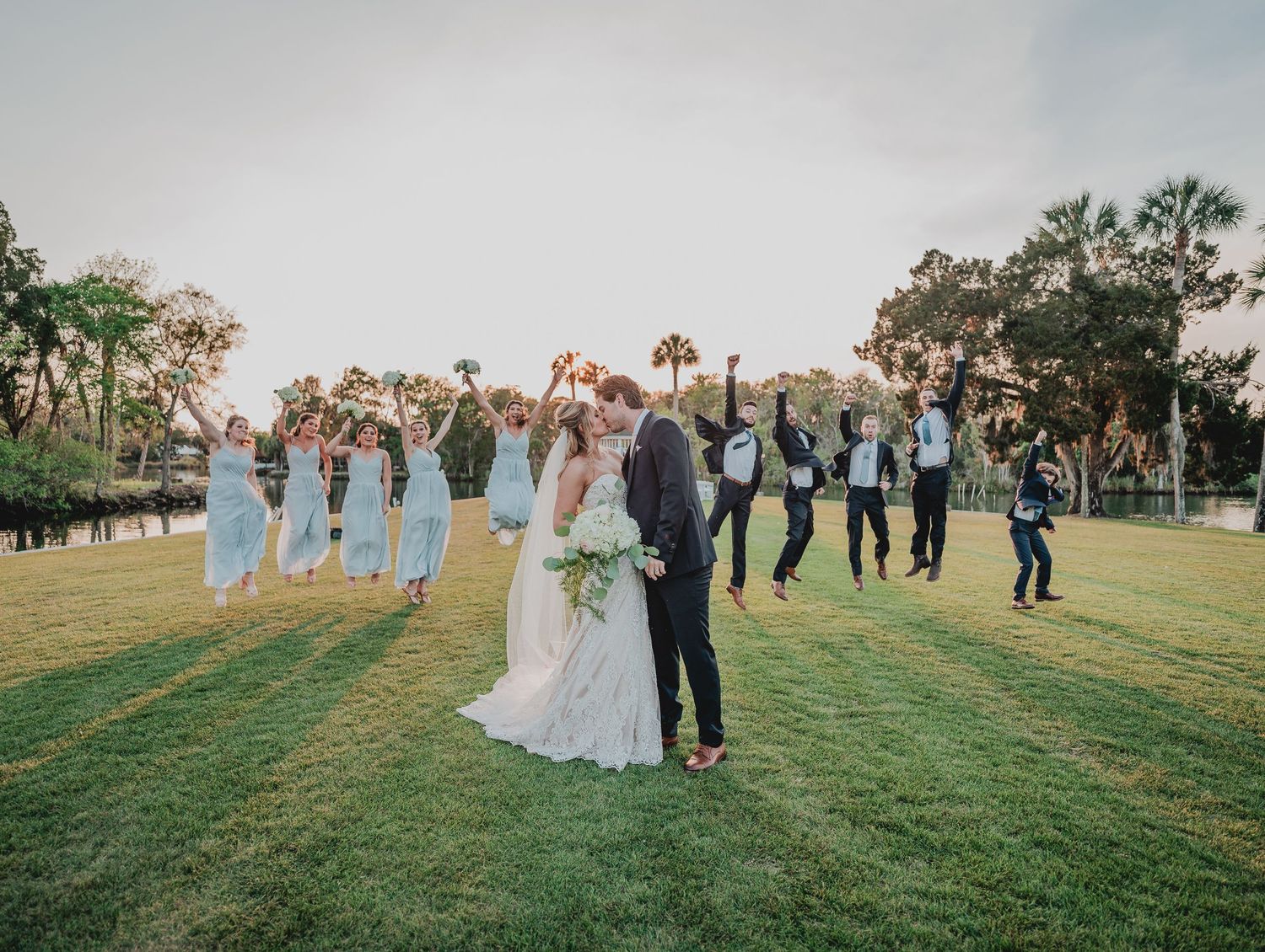 Wedding celebration on grassy lawn with celebratory group jump pose at sunset.