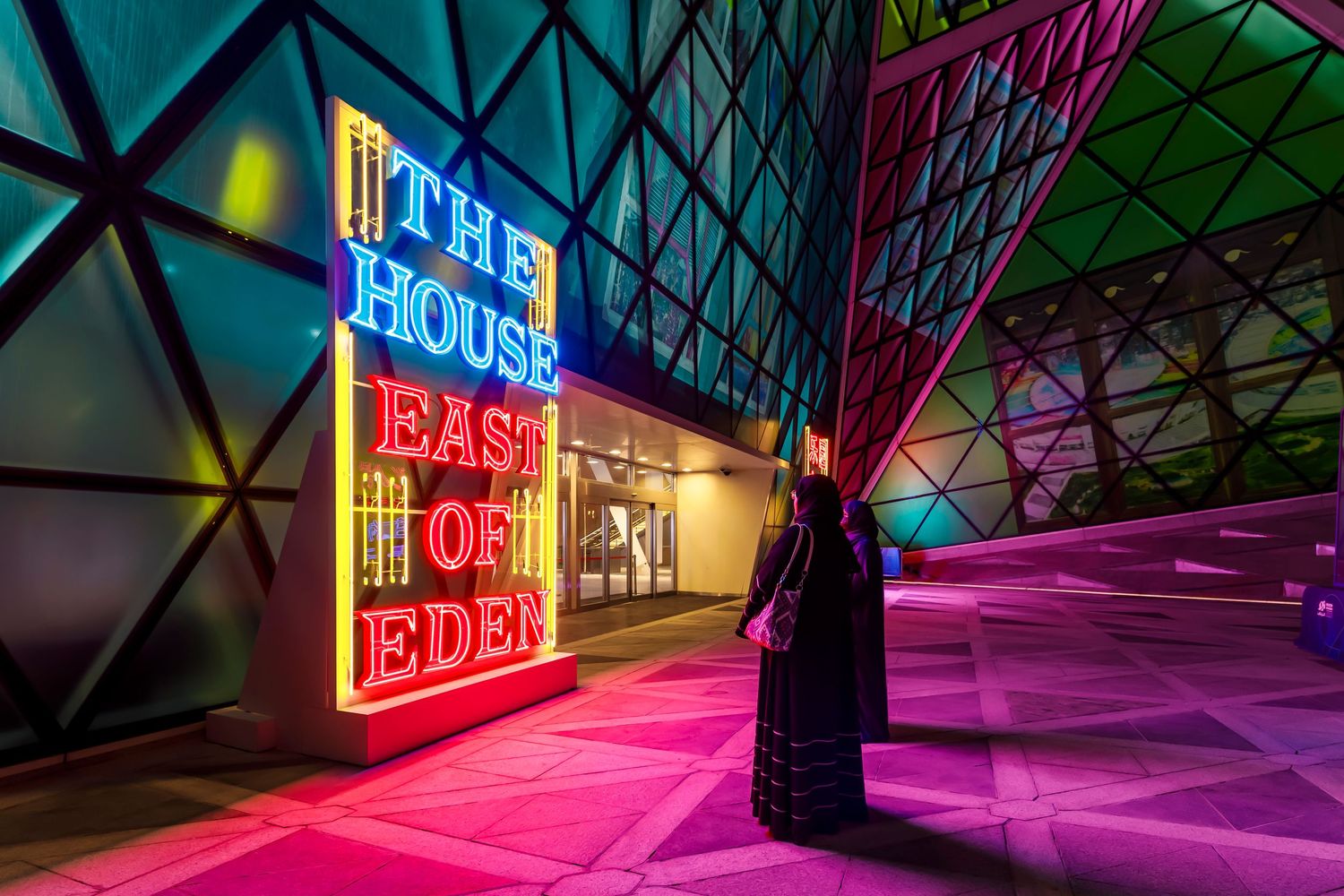 Neon signage glows in a geometric architectural space with colorful triangular patterns.