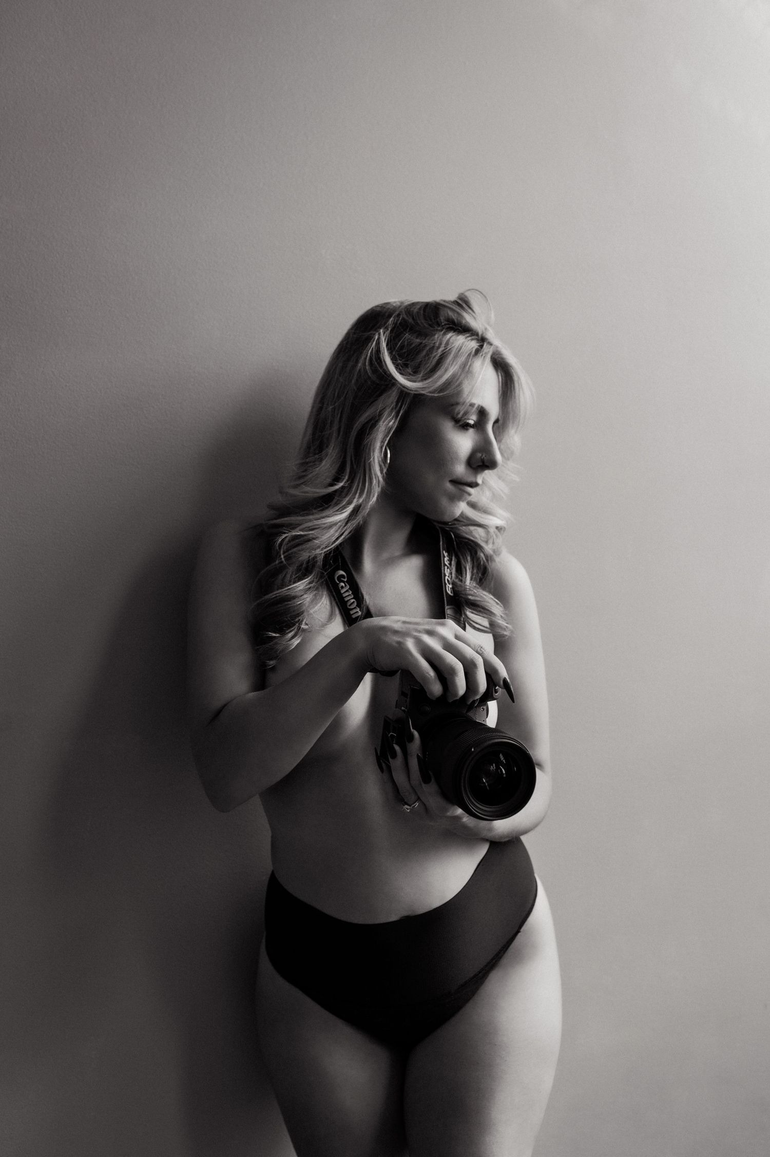 Black and white artistic boudoir photo of a woman in black underwear holding a camera against a plain wall.