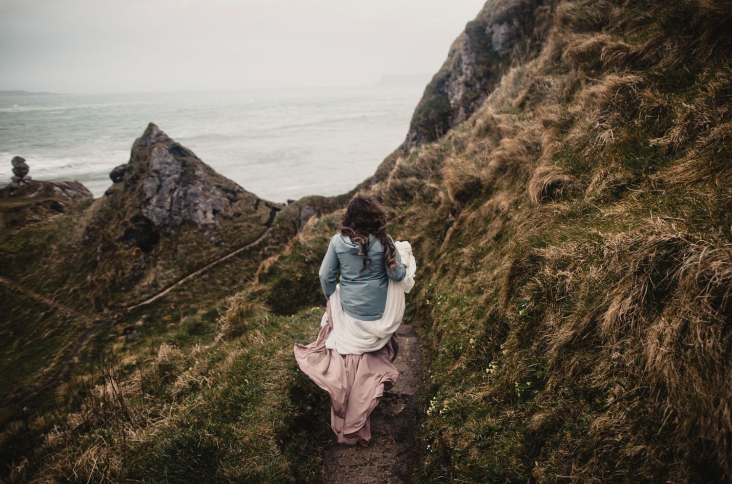 Person in casual wear sits on a clifftop overlooking dramatic coastal scenery.
