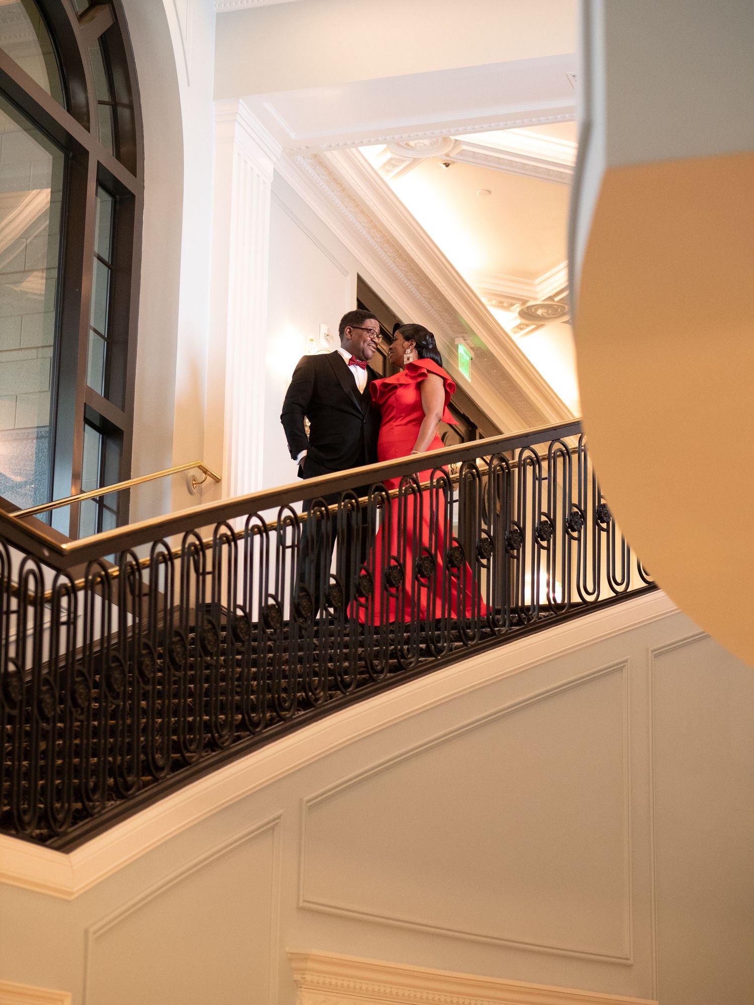 A couple in formal attire stands romantically on an elegant staircase with ornate iron railings.