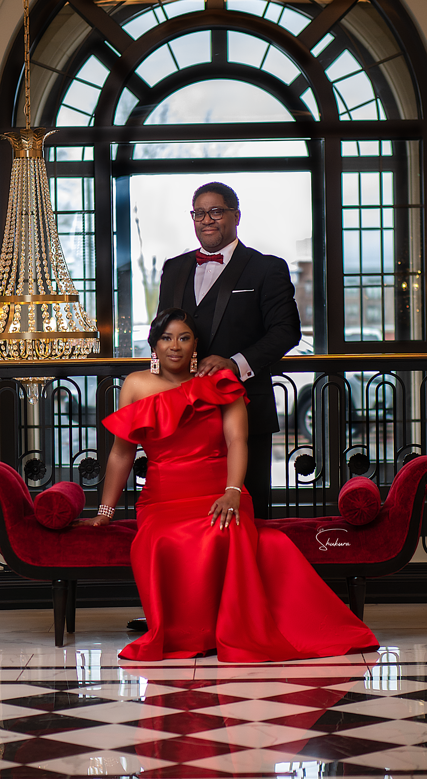 A couple poses elegantly on red velvet seating against an arched window and chandelier backdrop.