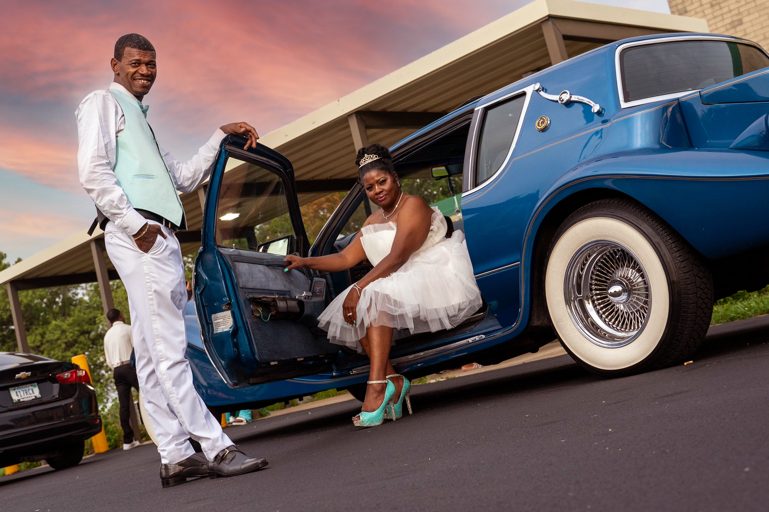 Stylish couple poses with a classic blue car at sunset.