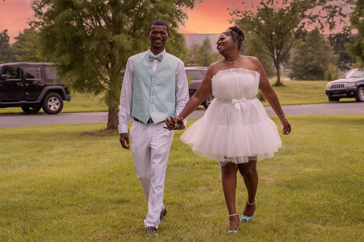 A couple walks hand in hand at sunset wearing white formal attire in a park setting.