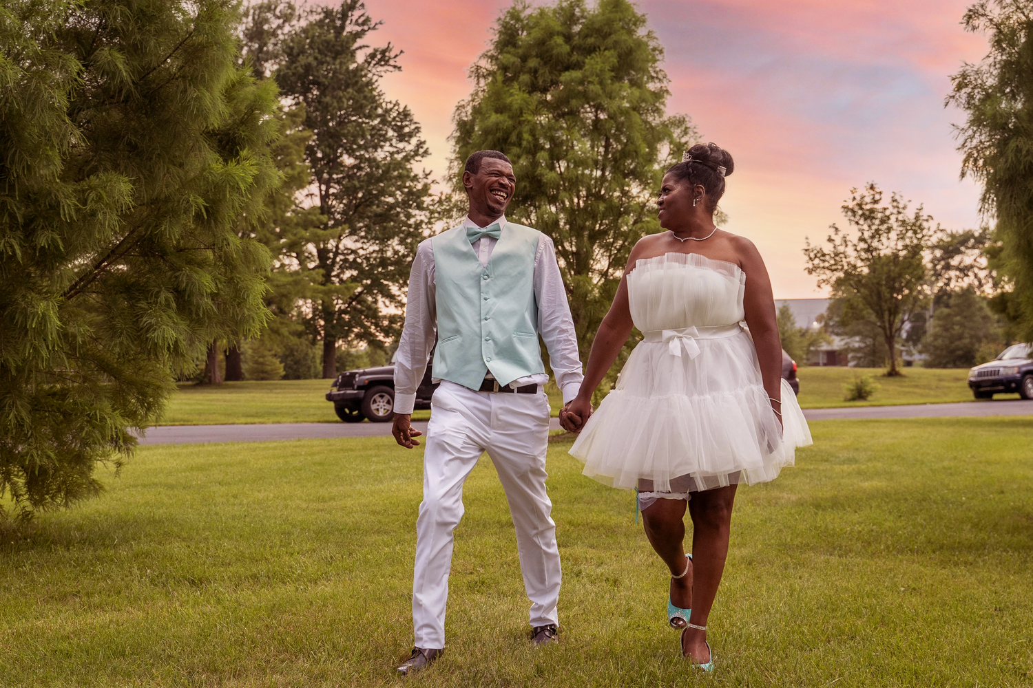 A couple strolls together at sunset wearing white formal outfits against a tree-lined backdrop.