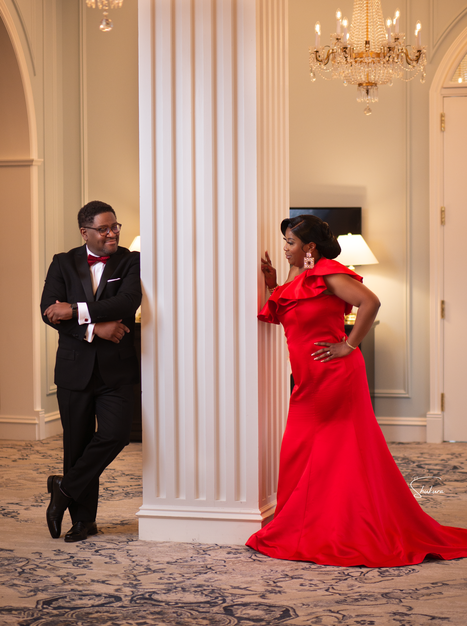 A couple in formal wear poses playfully in an elegant white room with a chandelier.
