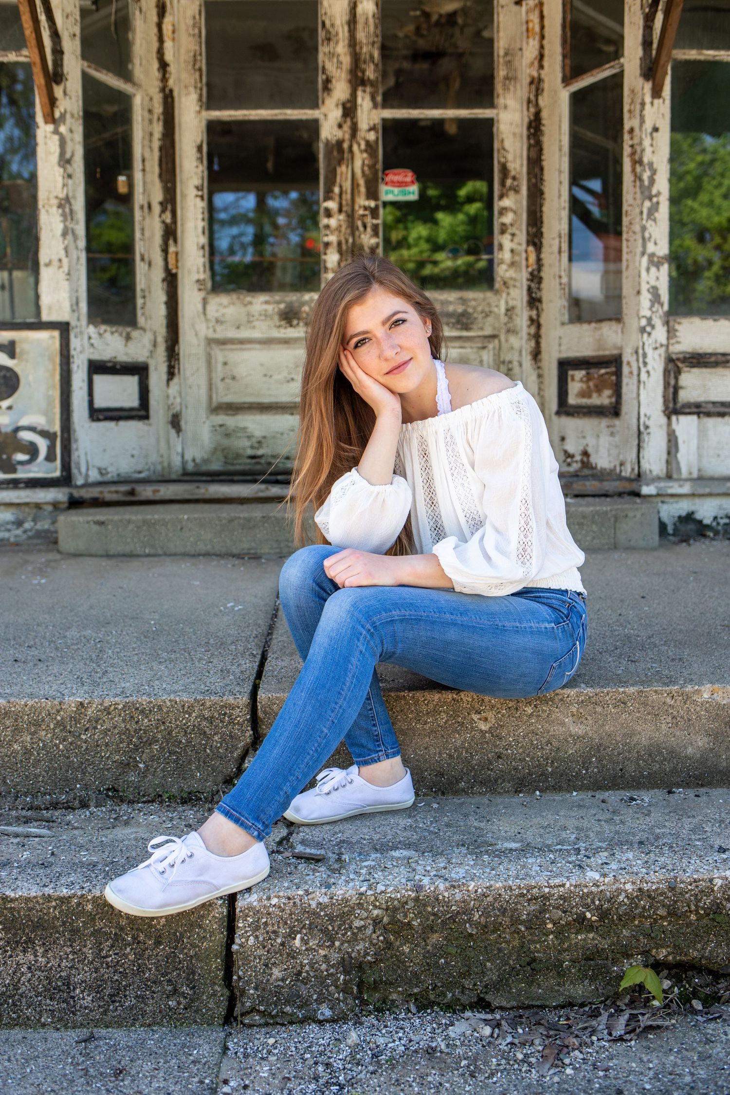 Young woman in white blouse and jeans sitting on steps of a rustic building.
