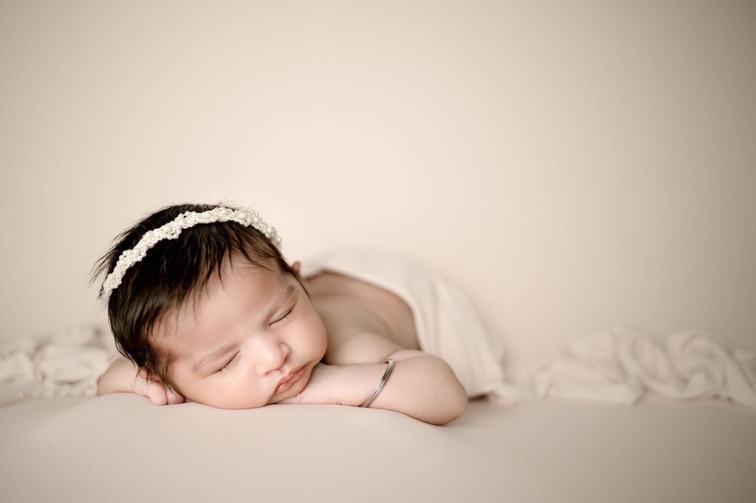 Serene newborn baby sleeping with white headband against soft beige background.