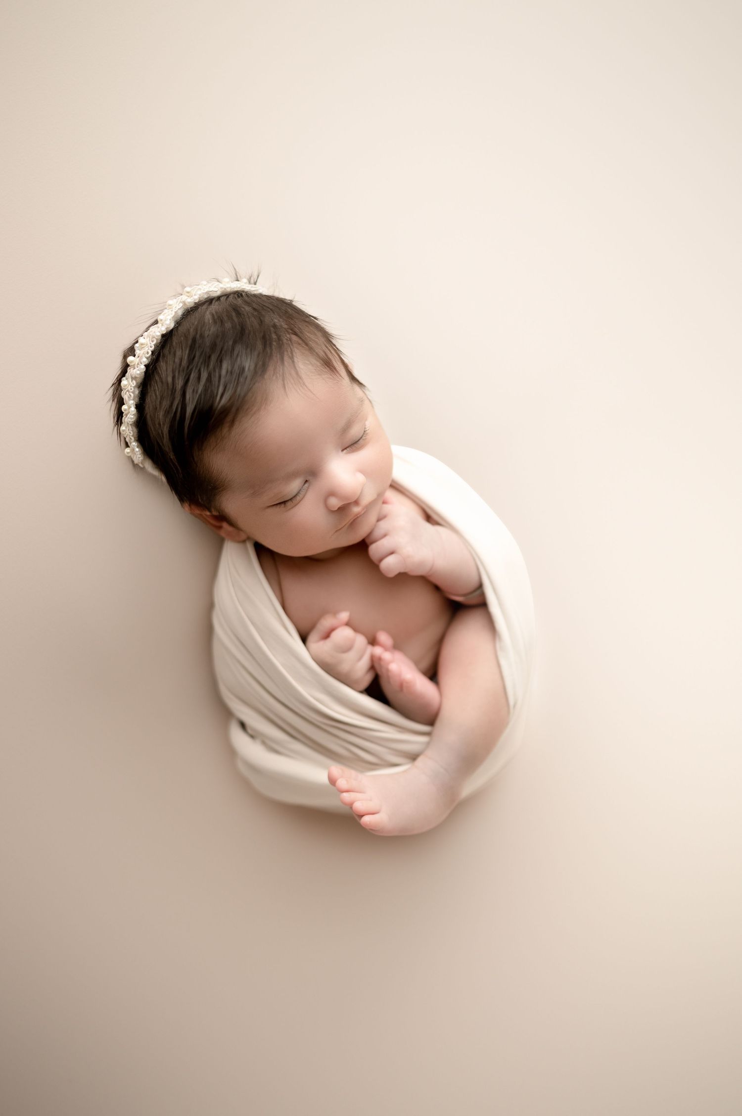 Sleeping newborn draped in white wrap with headband on beige background.
