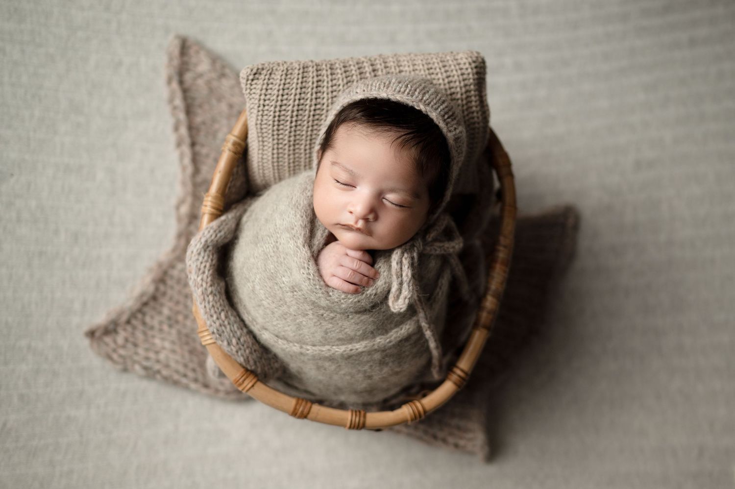 A newborn baby sleeping peacefully in a grey wrap inside a wooden basket on textured neutral background.