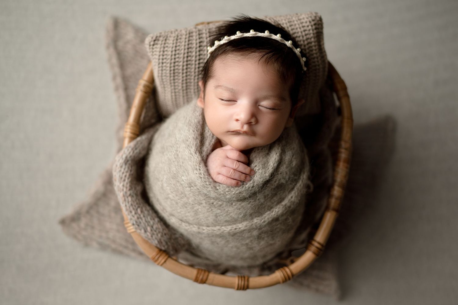 Sleeping newborn wrapped in gray swaddle positioned in wooden bowl with textured pillow.