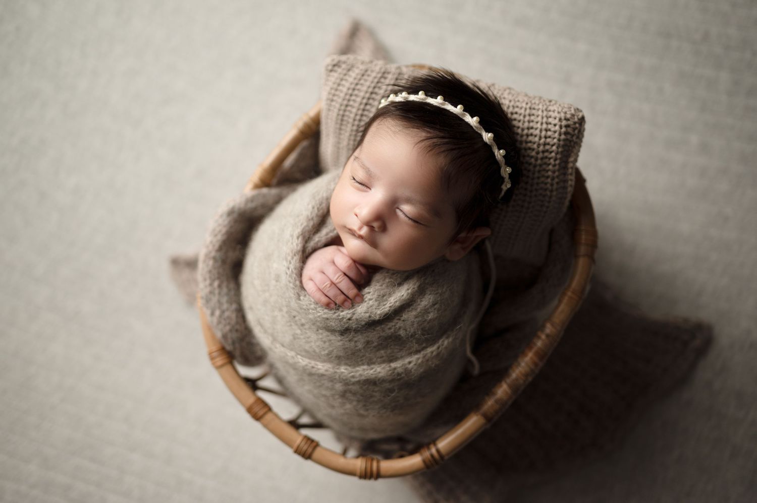 A newborn baby sleeping peacefully in a grey wrap inside a wooden basket on textured neutral background.