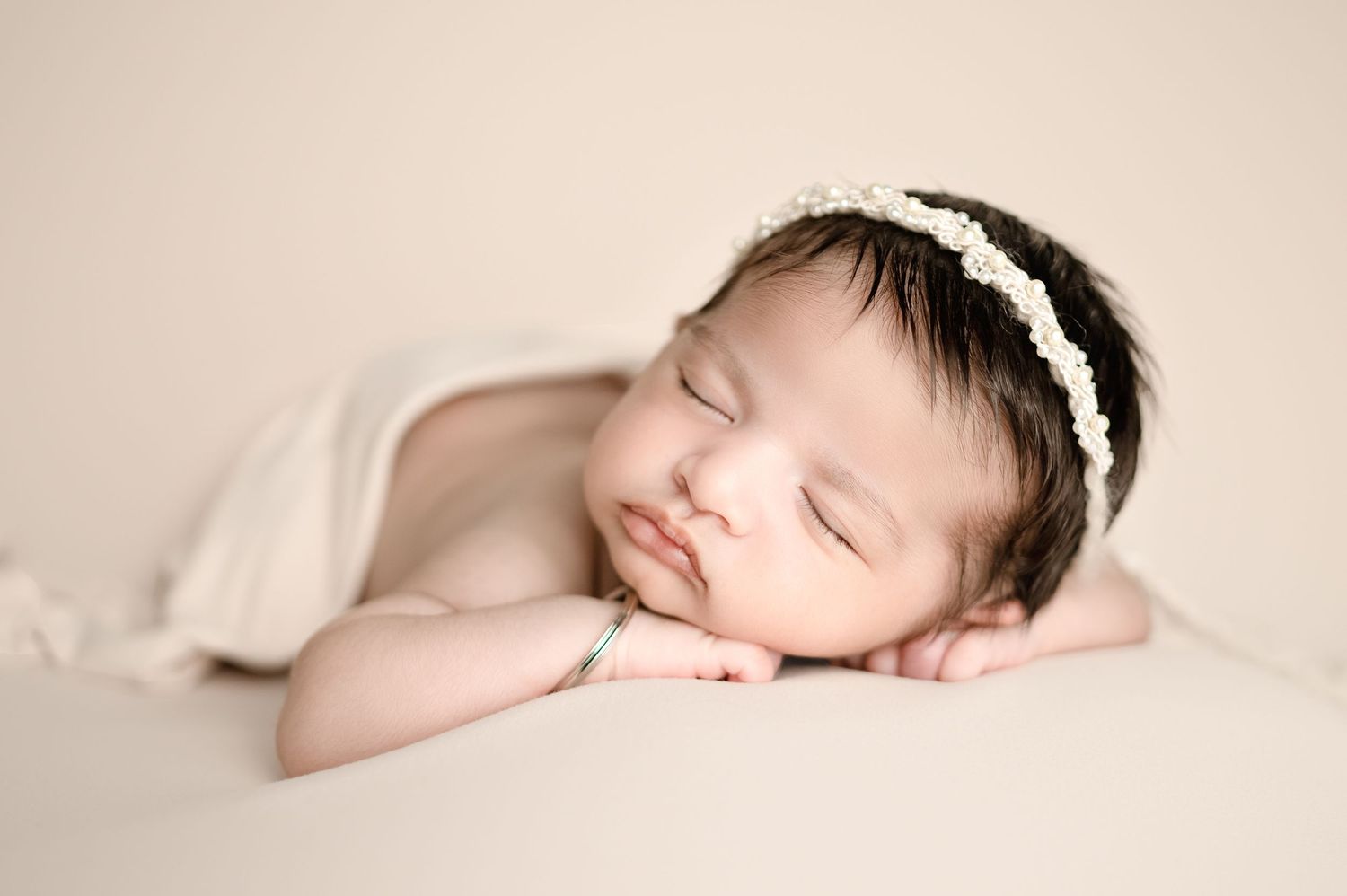 Peaceful sleeping newborn with lace headband resting on cream colored backdrop.
