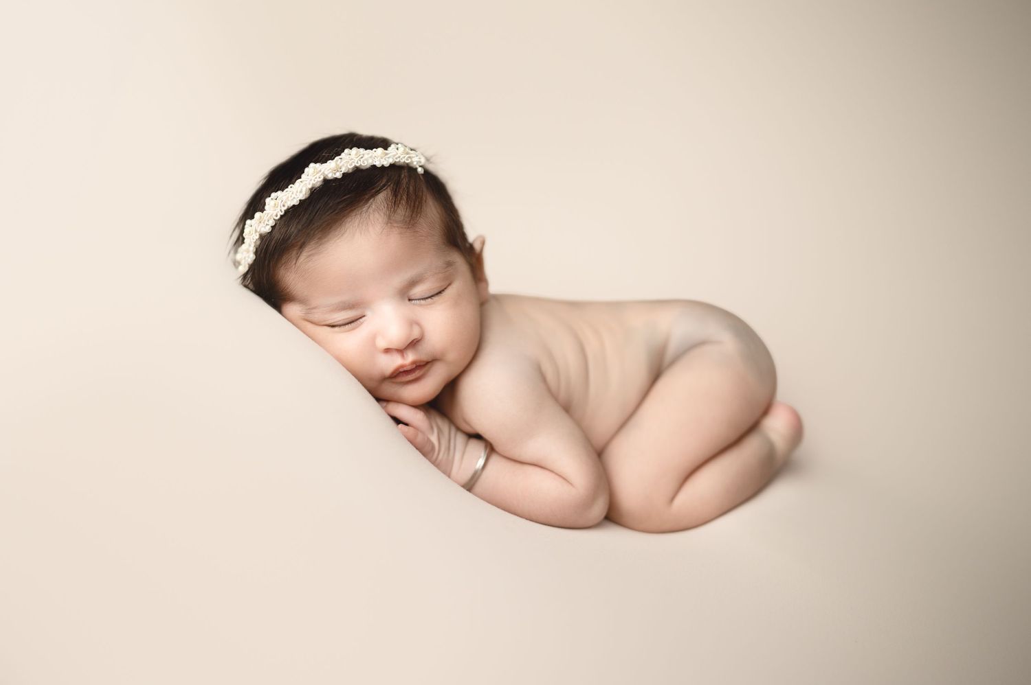 Sleeping newborn wearing delicate headband posed on cream background in studio setting.