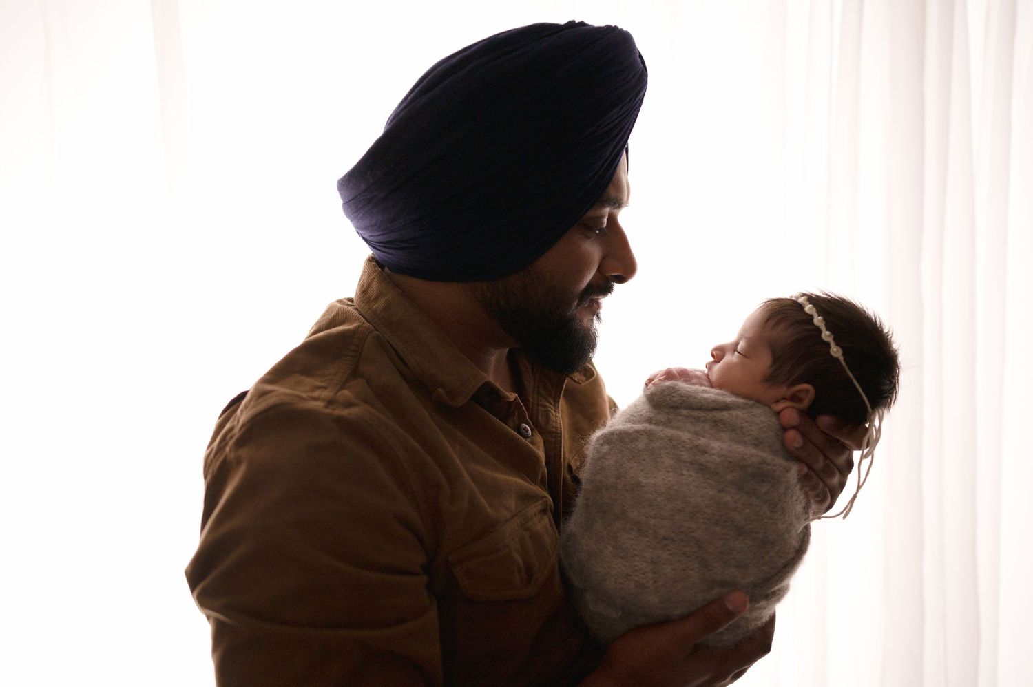Silhouette of parent in turban holding newborn baby against white window background.