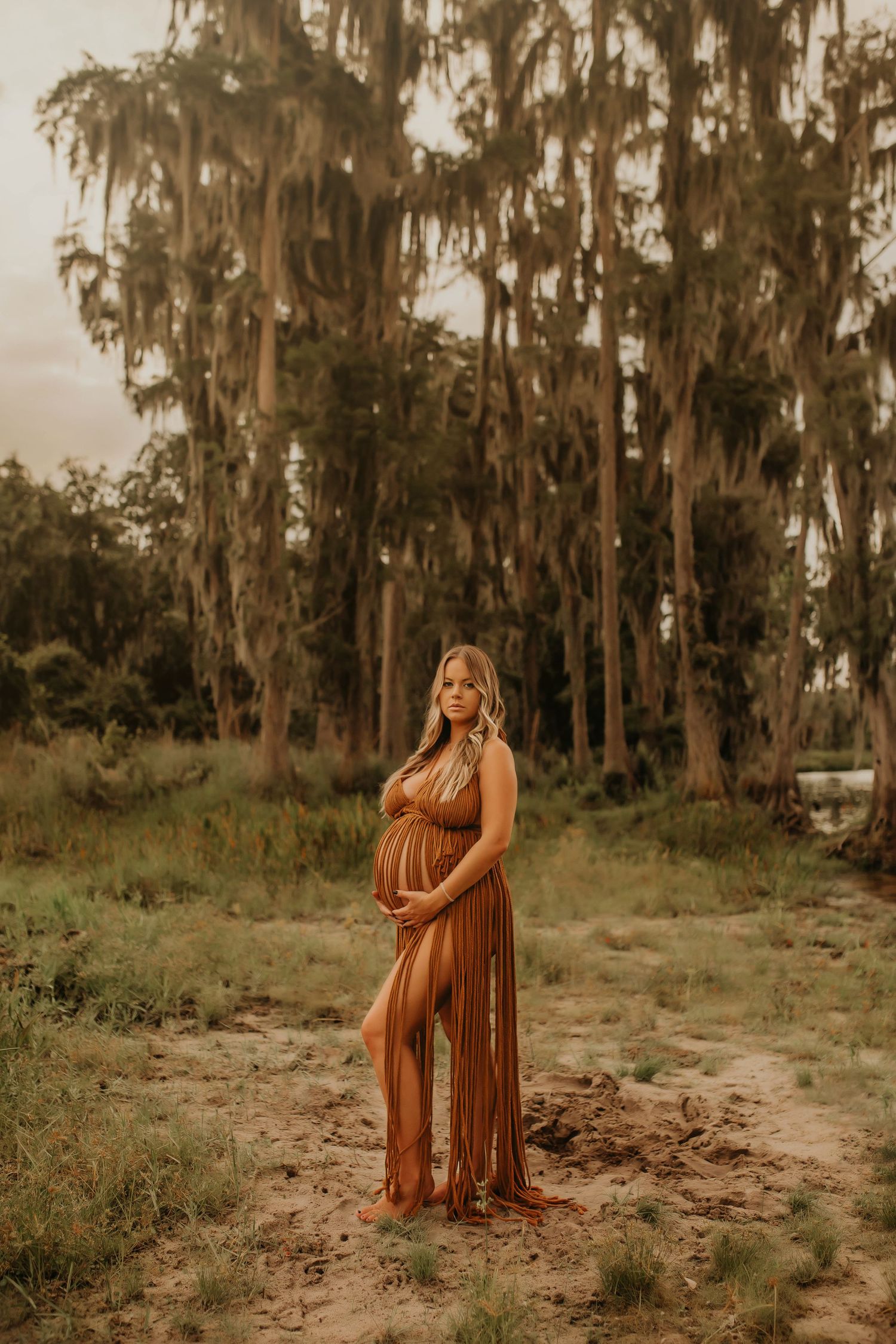 A woman in a brown maxi dress poses in front of Spanish moss-covered trees during golden hour.