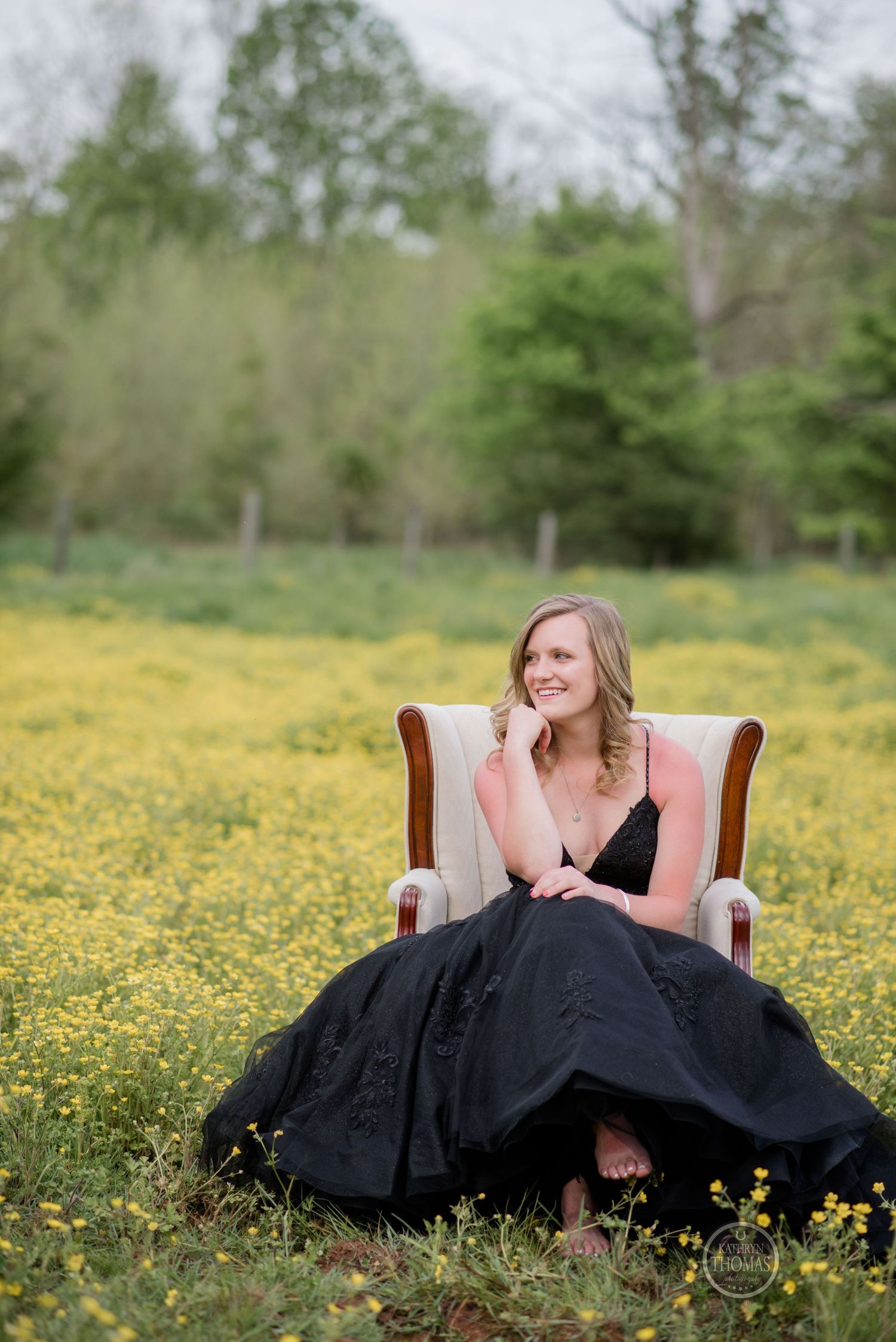 A figure in an elegant black gown poses in a vintage armchair amidst a field of yellow wildflowers.