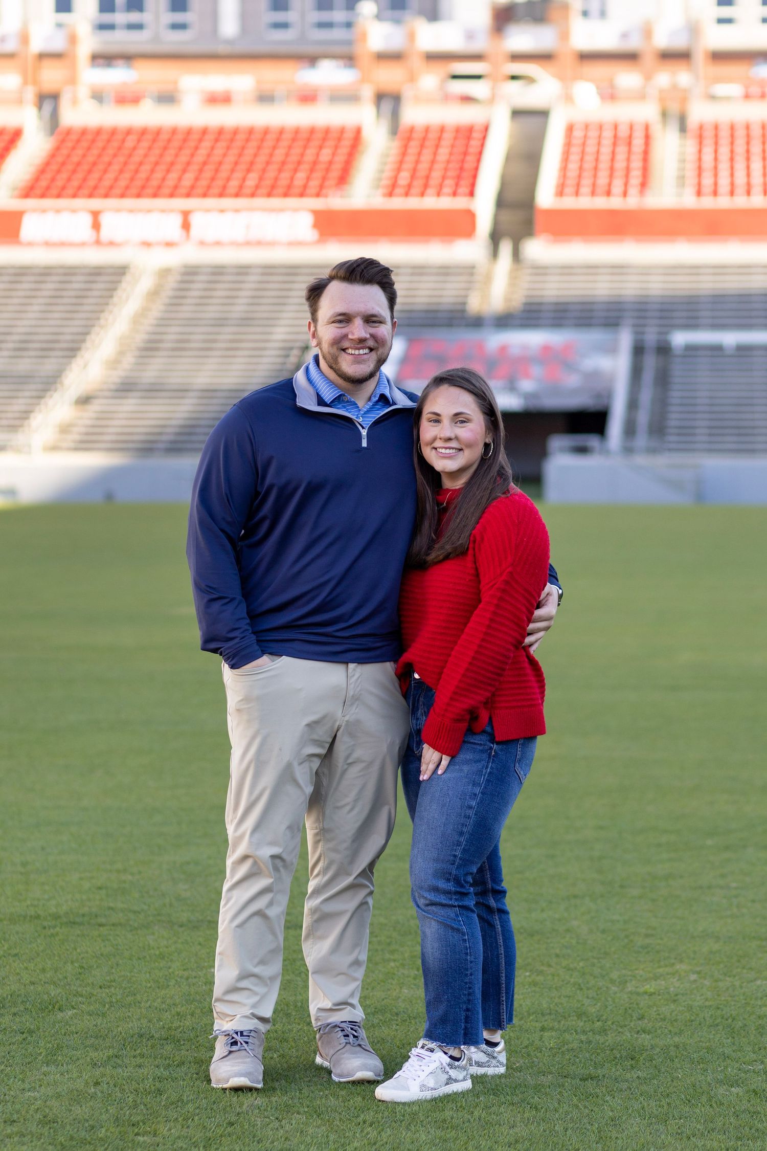 A couple dressed in a navy sweater and red sweater poses together on a football field in a stadium.