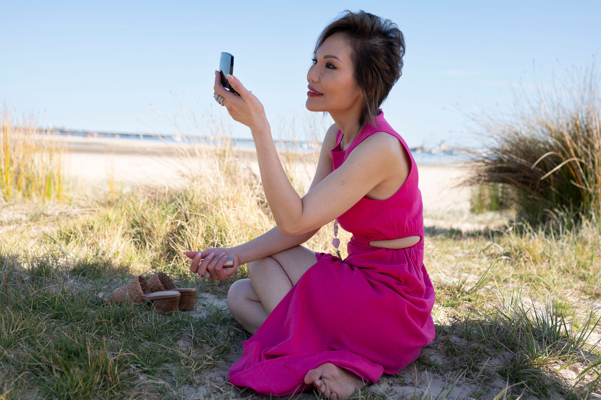 Woman in a pink dress sitting on grass taking a selfie at the beach.