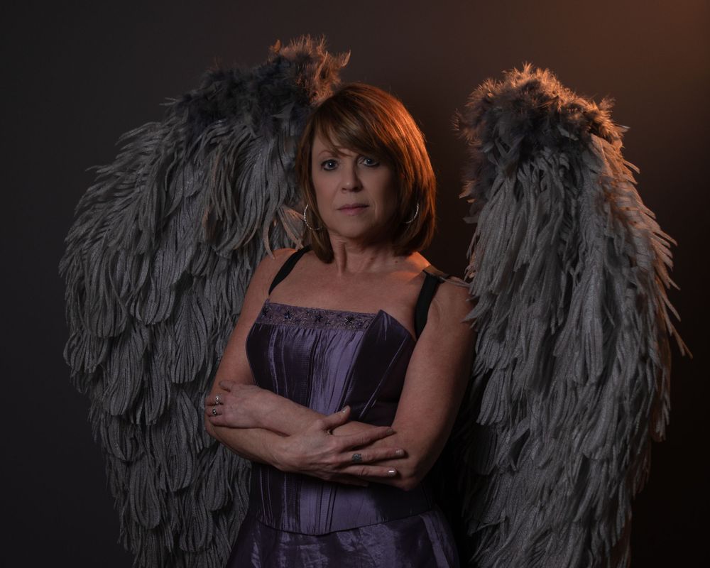 Dramatic portrait with large gray angel wings against dark background in purple dress.