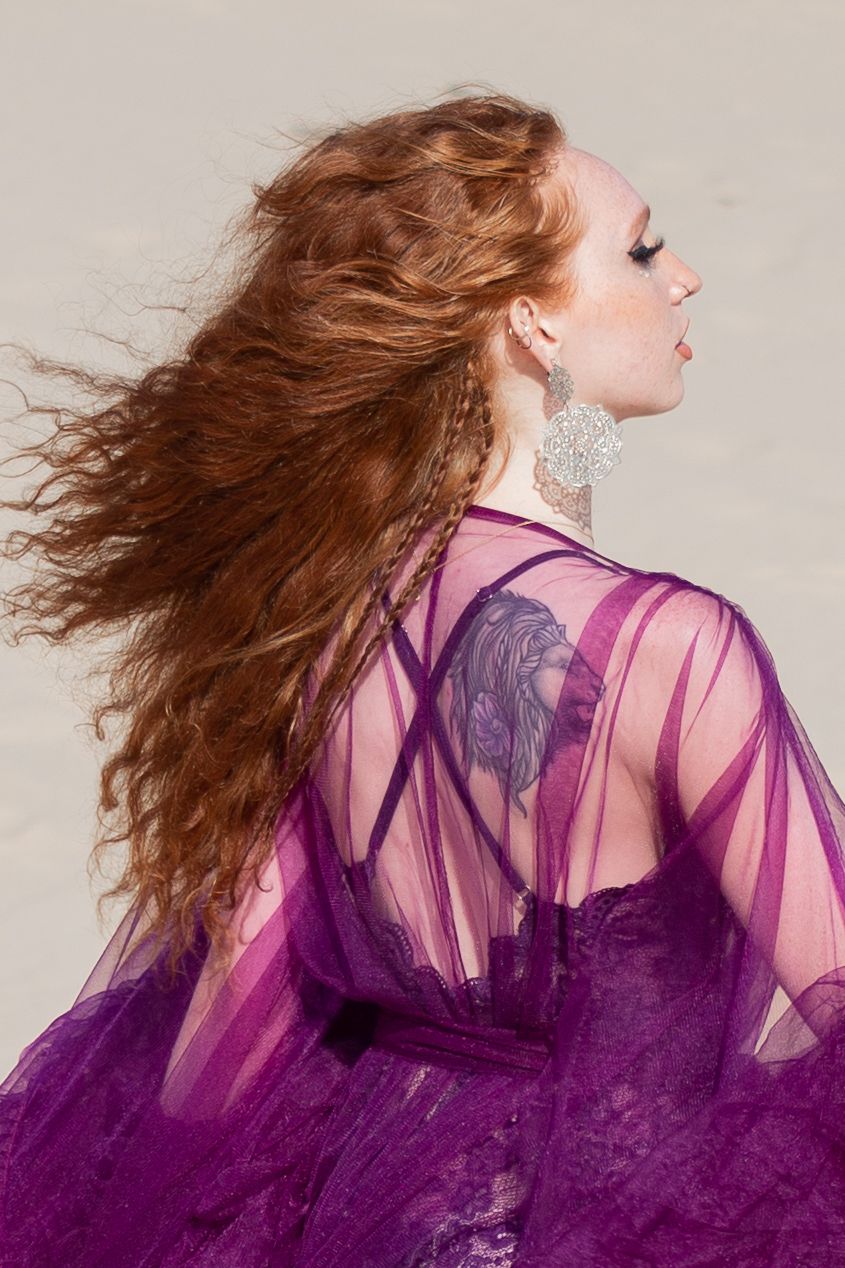 Dynamic fashion shot with flowing red hair and sheer purple gown against light background.