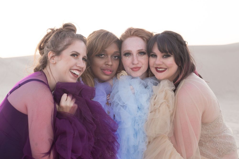 Four friends in pastel dresses share a joyful moment together against a light background.