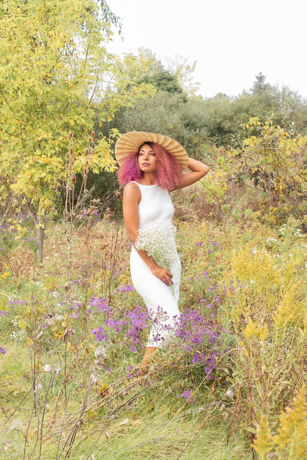 Someone in a white dress and wide-brimmed hat poses elegantly among wildflowers and tall grass.