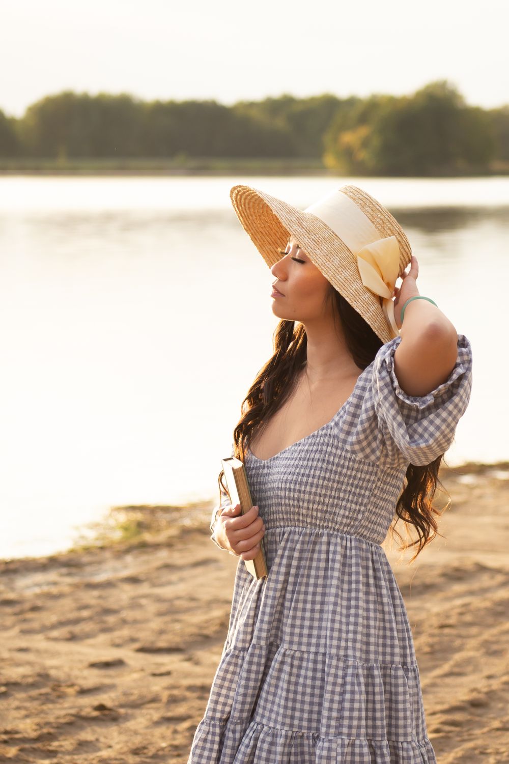 Romantic summer portrait in gingham dress and straw hat by lakeside at golden hour.