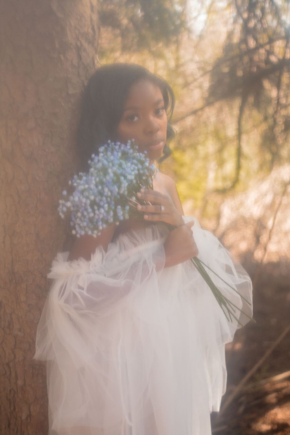 Ethereal portrait holding white flowers in flowing white dress against sunlit background.