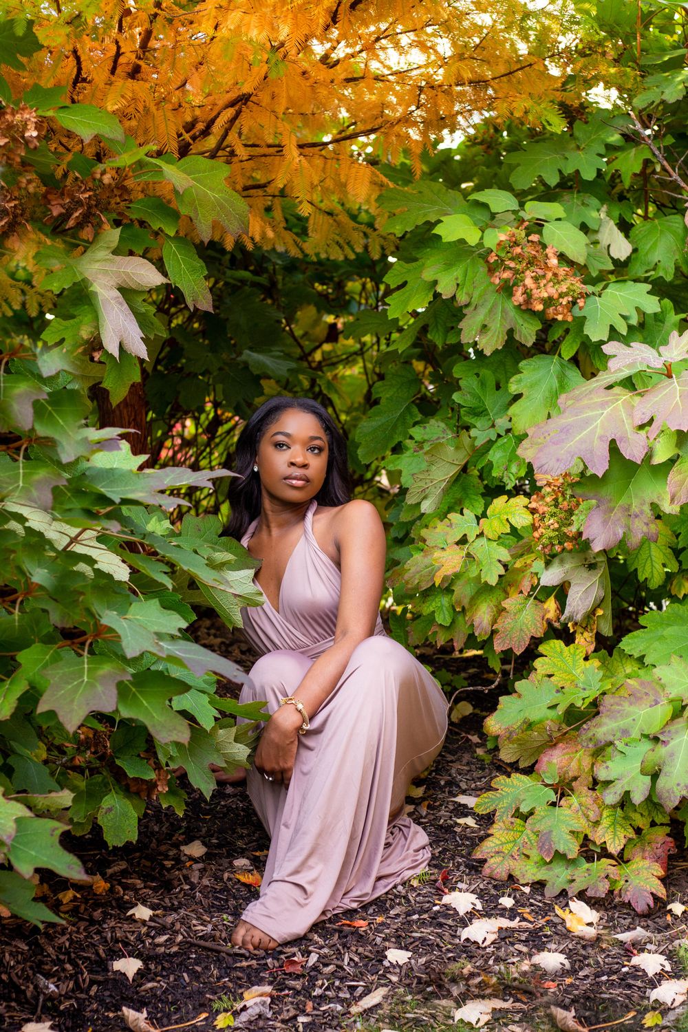 Elegant portrait in a blush pink gown against autumn foliage with golden and green leaves.