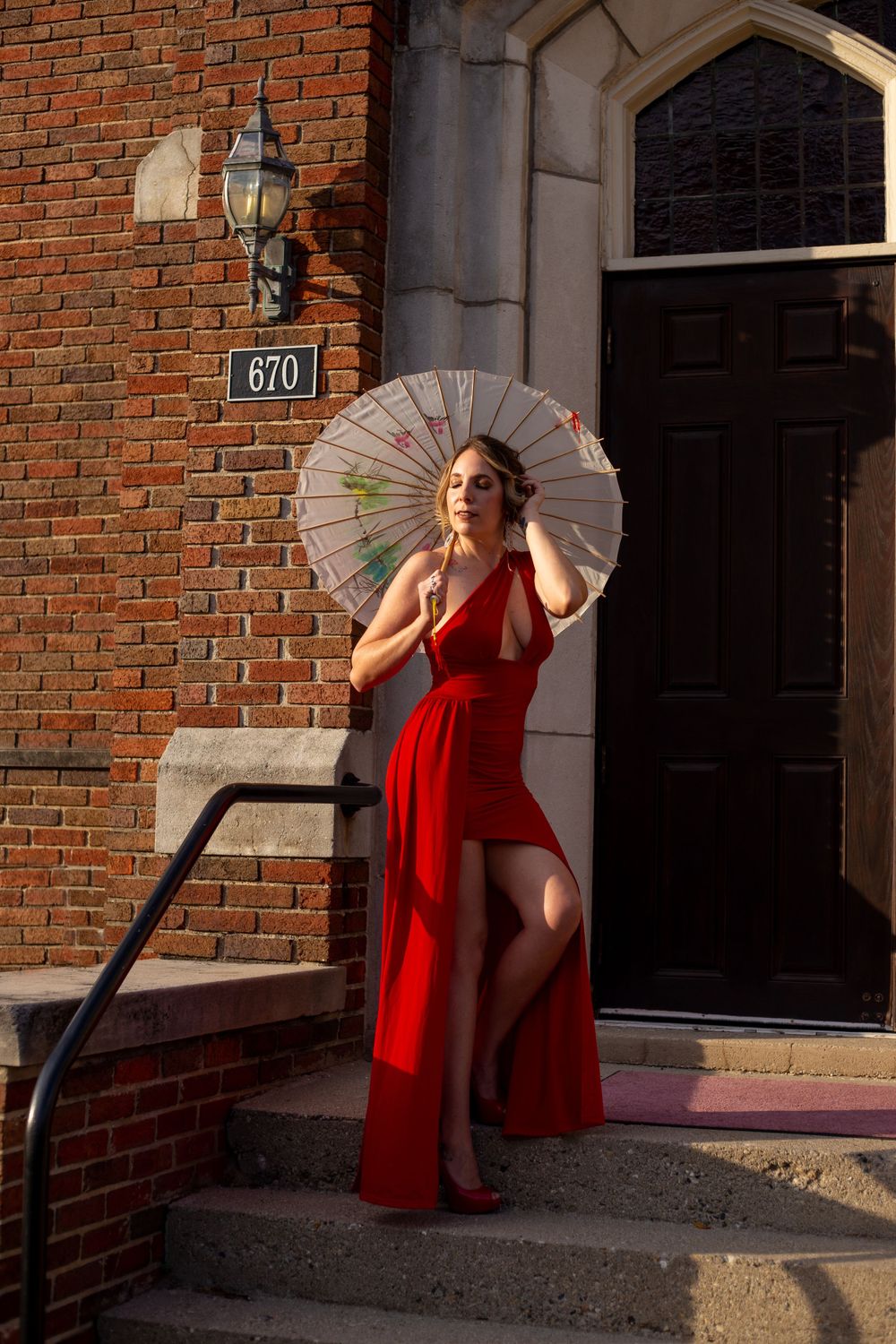 Dramatic fashion pose with parasol against brick building entrance in red flowing dress.