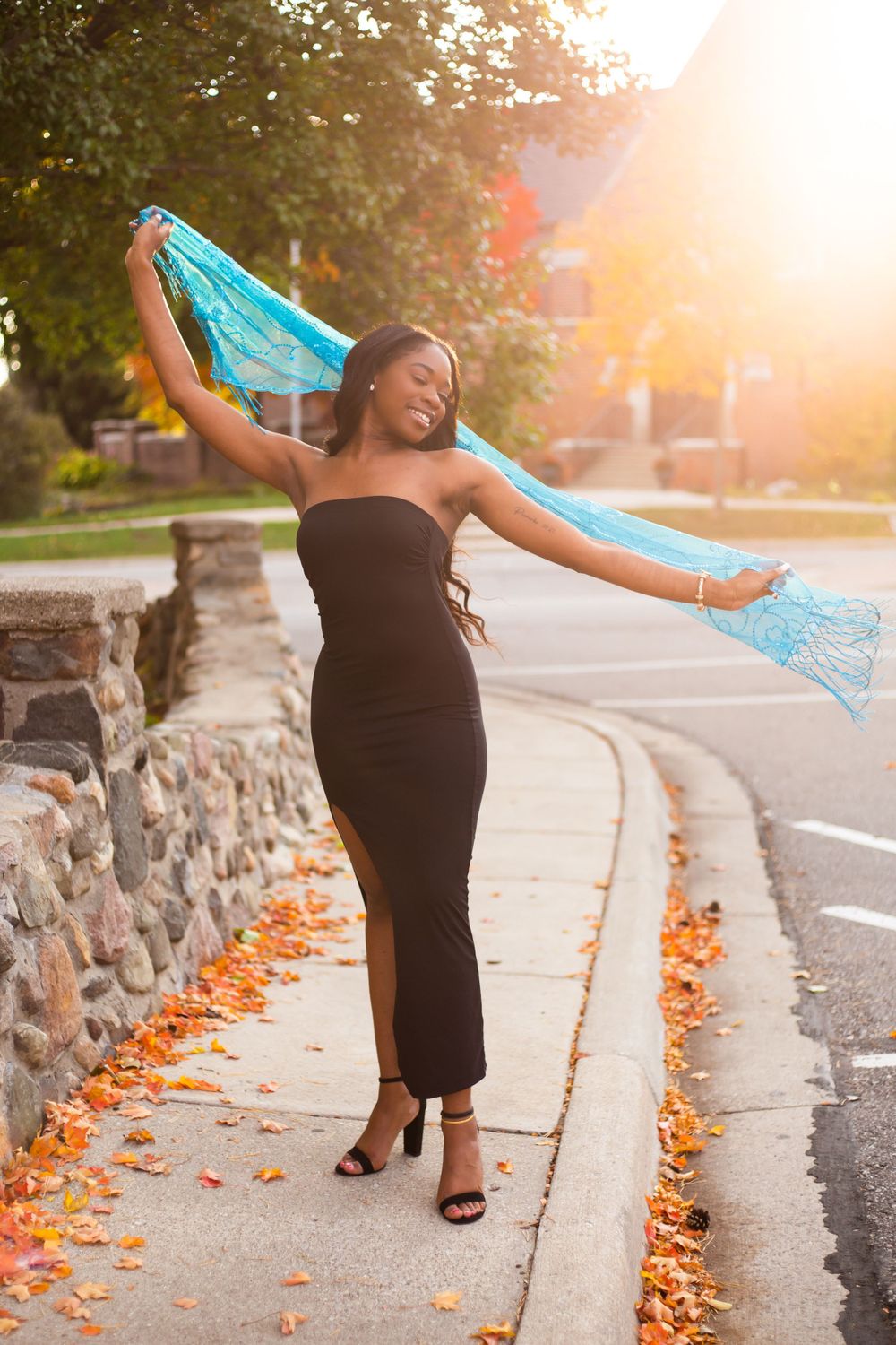Fashion portrait with flowing blue scarf at sunset against stone wall with autumn leaves.